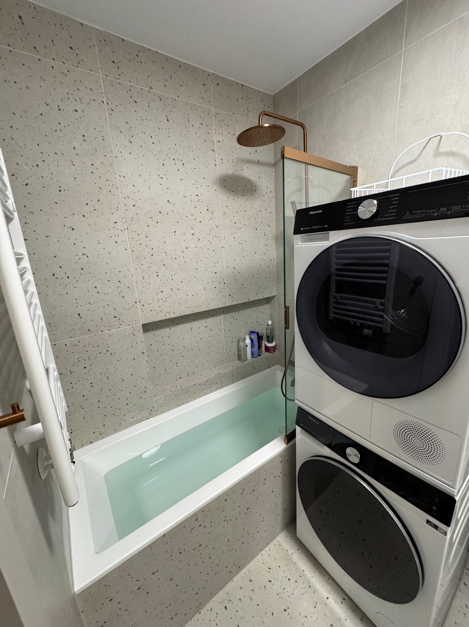 Nowoczesna łazienka z wanną wypełnioną wodą, szklanym parawanem, deszczownicą i pralko-suszarką Hisense w jasnej tonacji z mozaikową fakturą płytek.