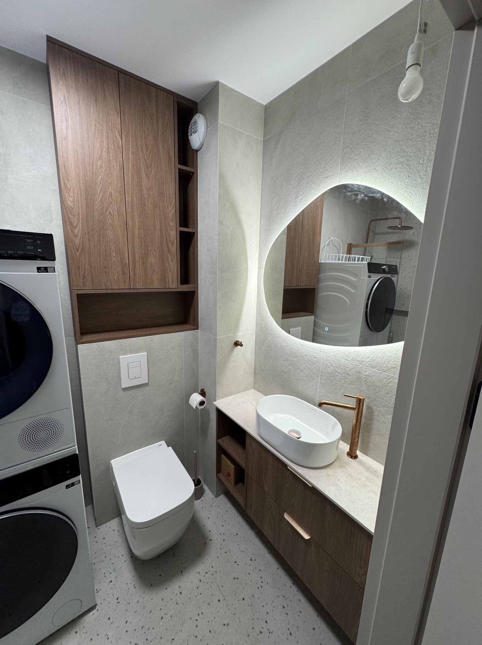 Nowoczesna łazienka z drewnianymi szafkami, owalnym lustrem z podświetleniem LED, białą umywalką i toaletą. Pralka i suszarka w rogu. Minimalistyczny design.