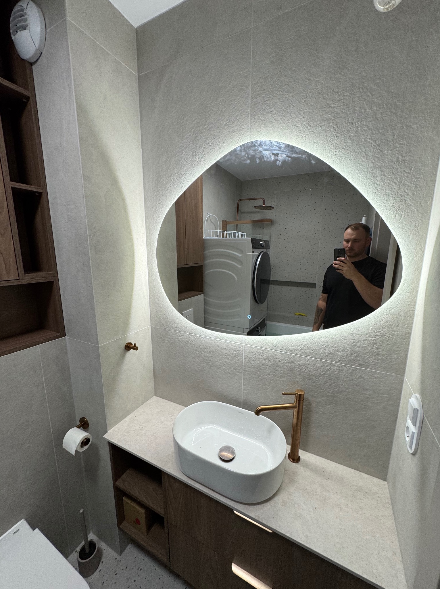 Nowoczesna łazienka z umywalką nablatową, baterią w kolorze miedzi i podświetlanym lustrem o nieregularnym kształcie. W tle widoczna pralka i kabina prysznicowa.