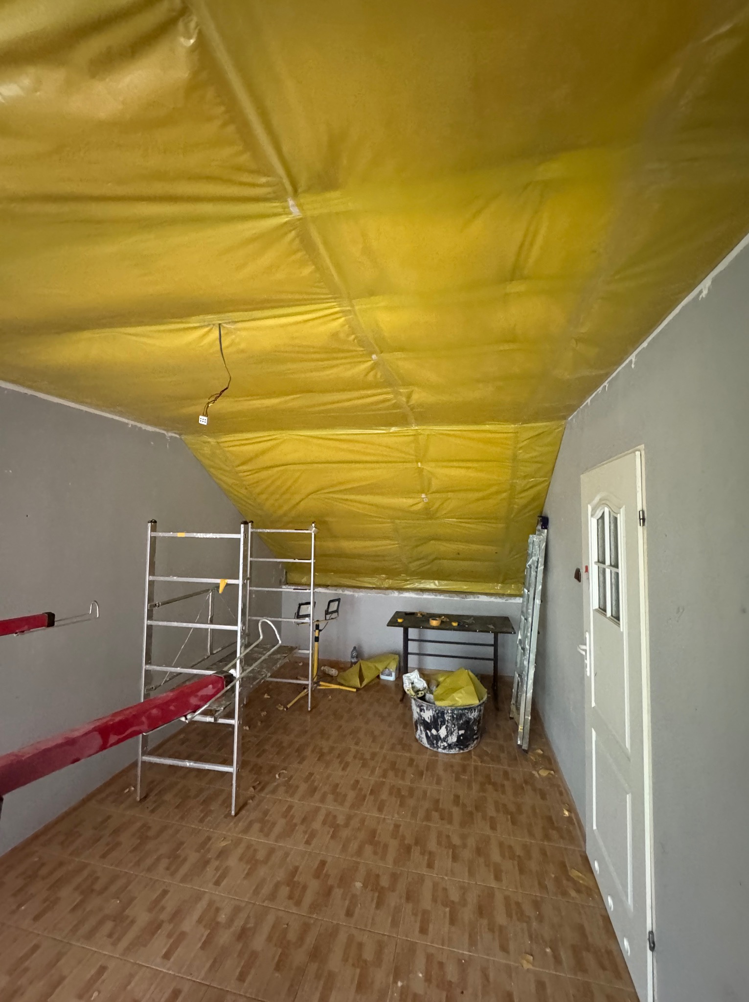 Pokój w trakcie prac wykończeniowych: żółta folia na suficie, rusztowanie, drabina, wiadro z farbą. Widoczne przygotowania do dalszych prac remontowych.
