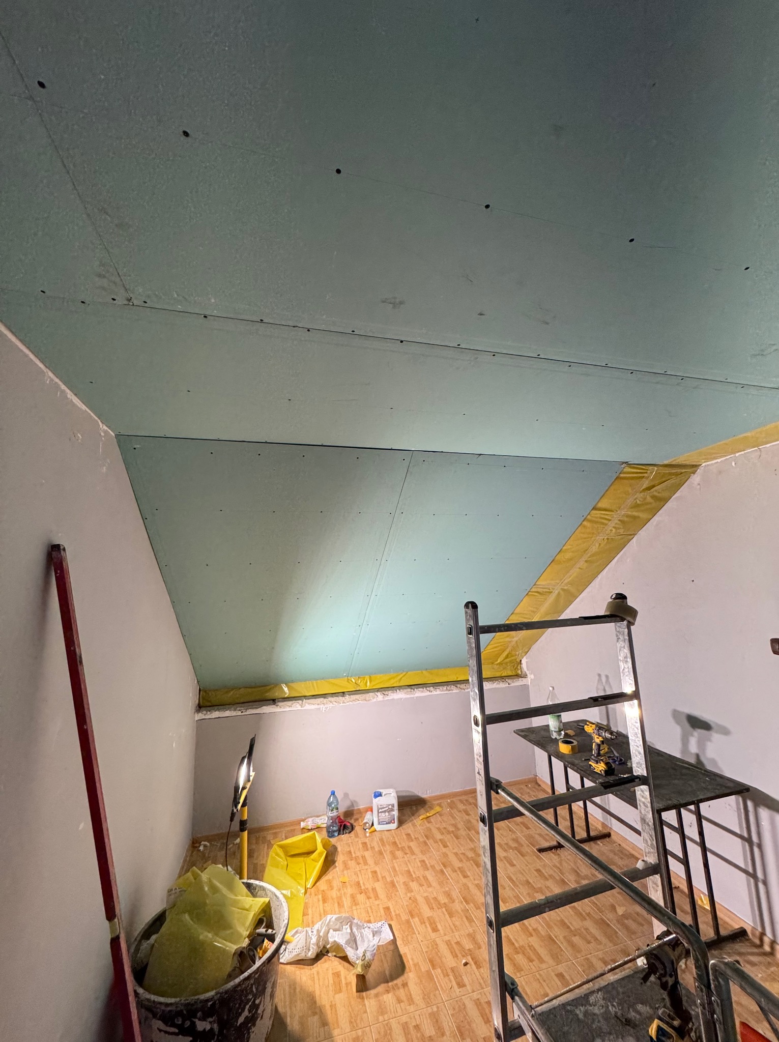 Montaż zielonych płyt karton-gips na skośnym suficie poddasza, widoczna drabina, narzędzia i oświetlenie budowlane. Żółta izolacja na styku ściany i sufitu.