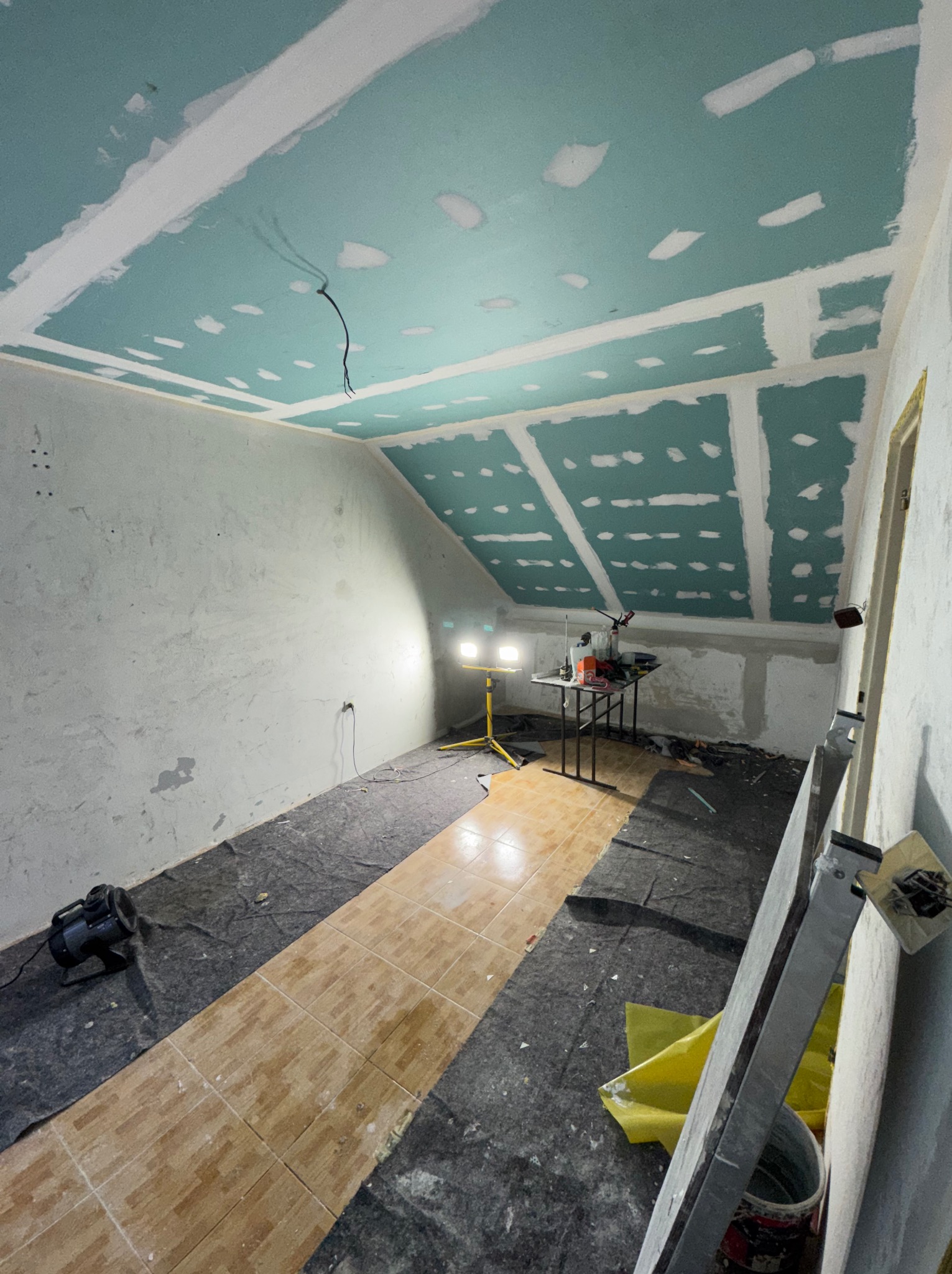 Poddasze w trakcie montażu płyt karton-gips na skośnym suficie. Widoczne narzędzia, lampa oświetleniowa i zabezpieczona podłoga. Etap szpachlowania łączeń.