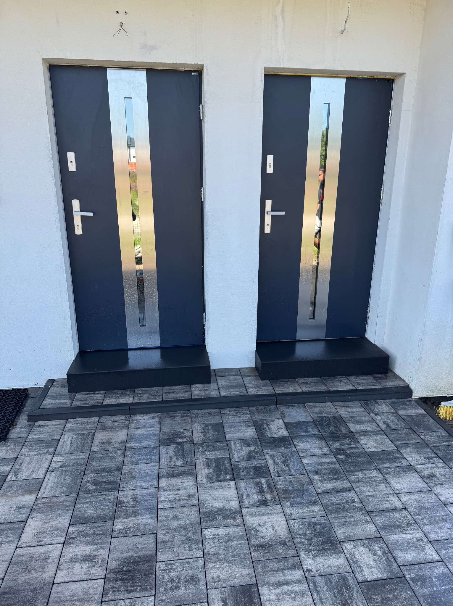 Dwa nowoczesne, grafitowe drzwi wejściowe z pionowymi, srebrnymi wstawkami, prowadzące do domu z szarą kostką brukową. Widoczne ślady po montażu nad drzwiami.