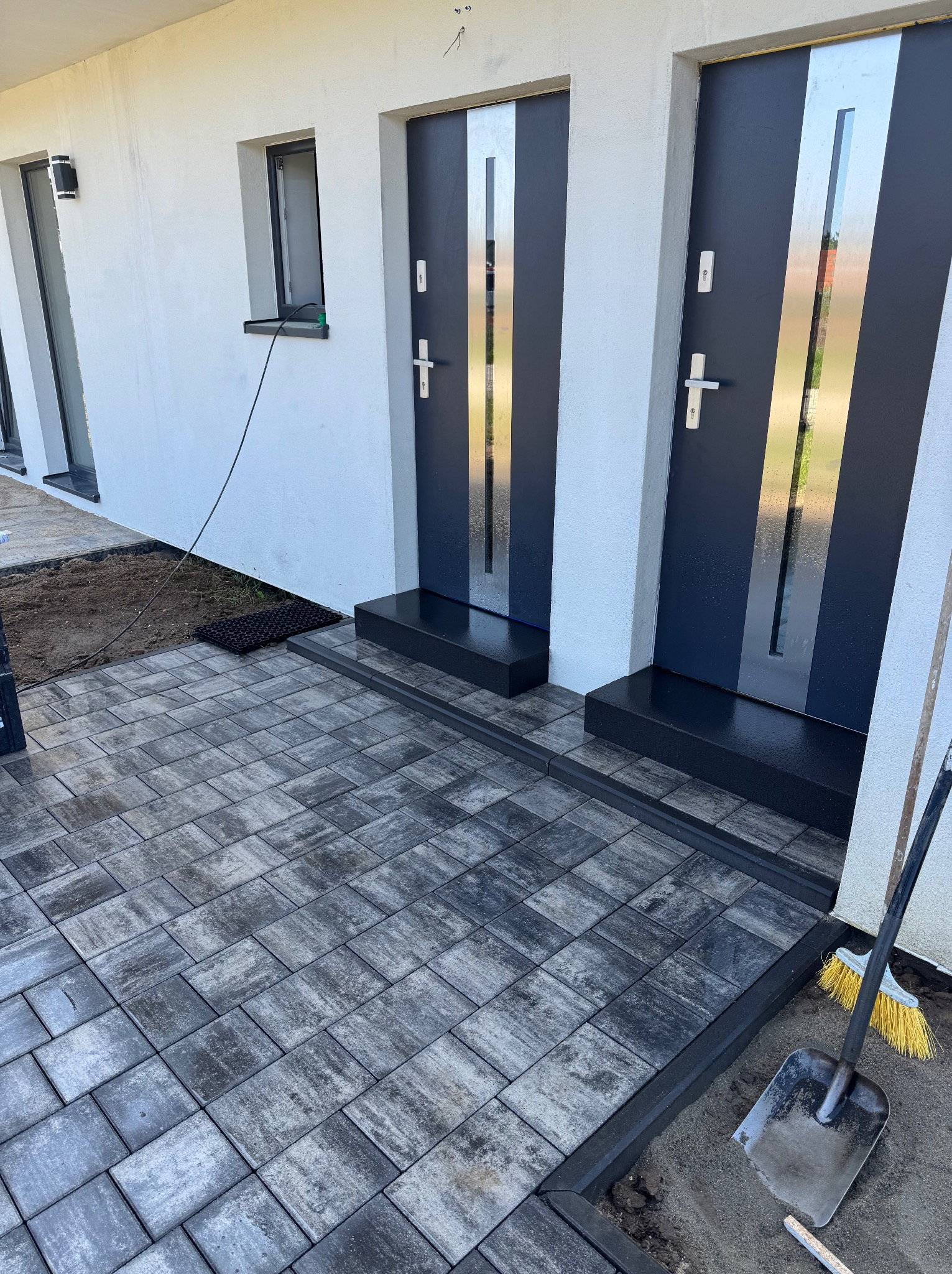 Ułożona kostka brukowa przed wejściem do budynku z dwoma parami drzwi. Widoczne narzędzia: łopata i miotła. Szara kostka, czarne obramowanie.