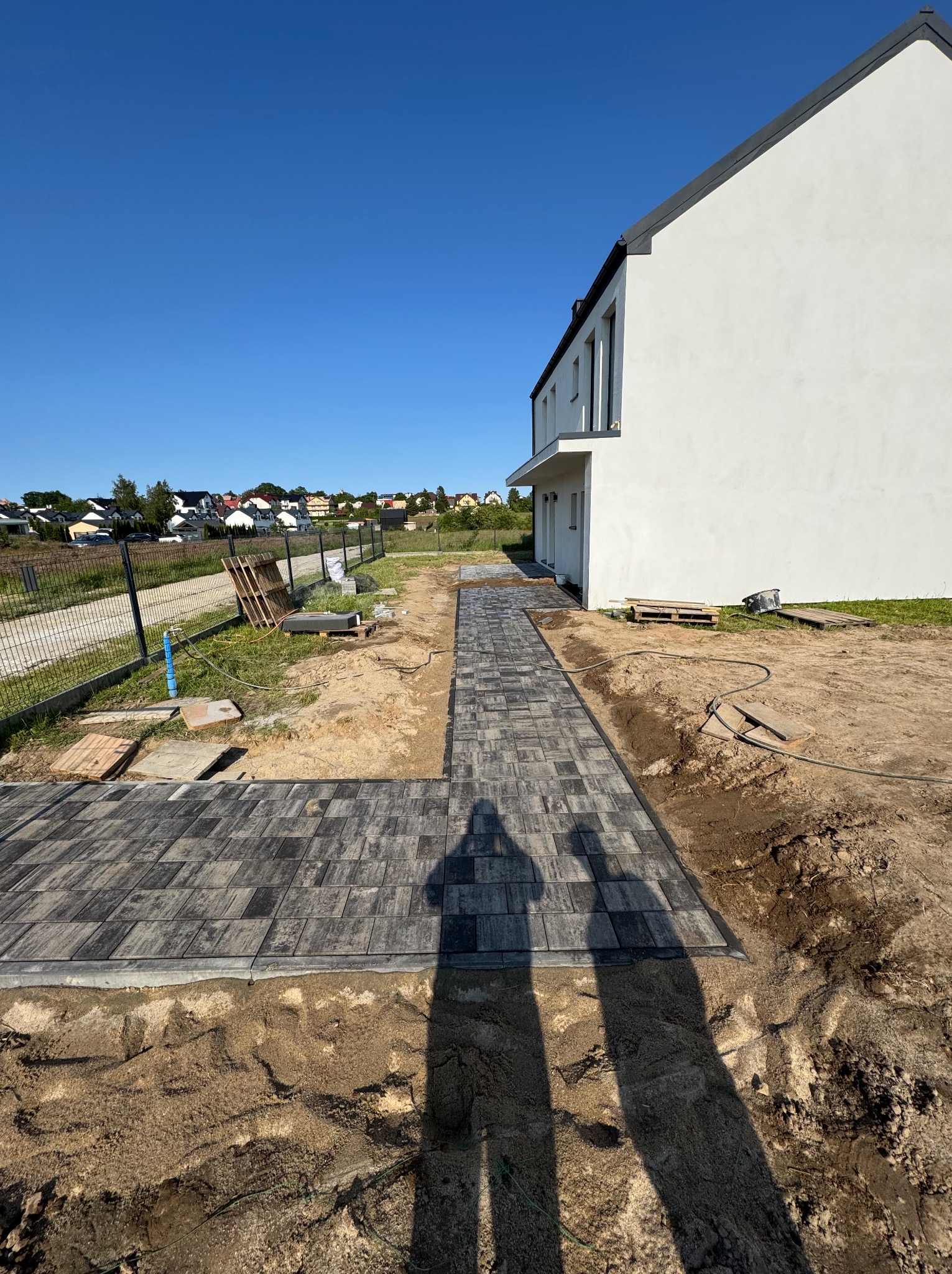 Układanie szarej kostki brukowej na posesji domu jednorodzinnego w słoneczny dzień. Widoczne narzędzia i cień osoby wykonującej prace.