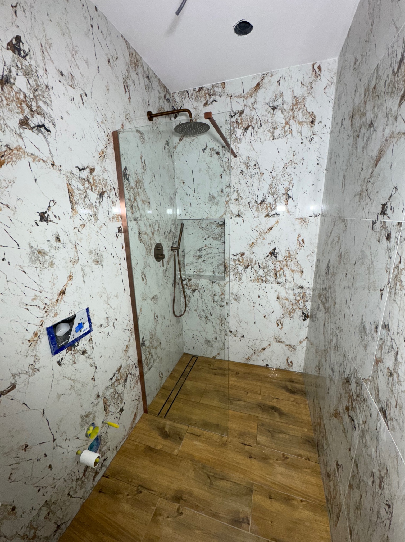 Wykończona kabina prysznicowa z marmurowymi płytkami i drewnopodobną podłogą. Widoczne elementy armatury w kolorze miedzi i przygotowane miejsce na toaletę.