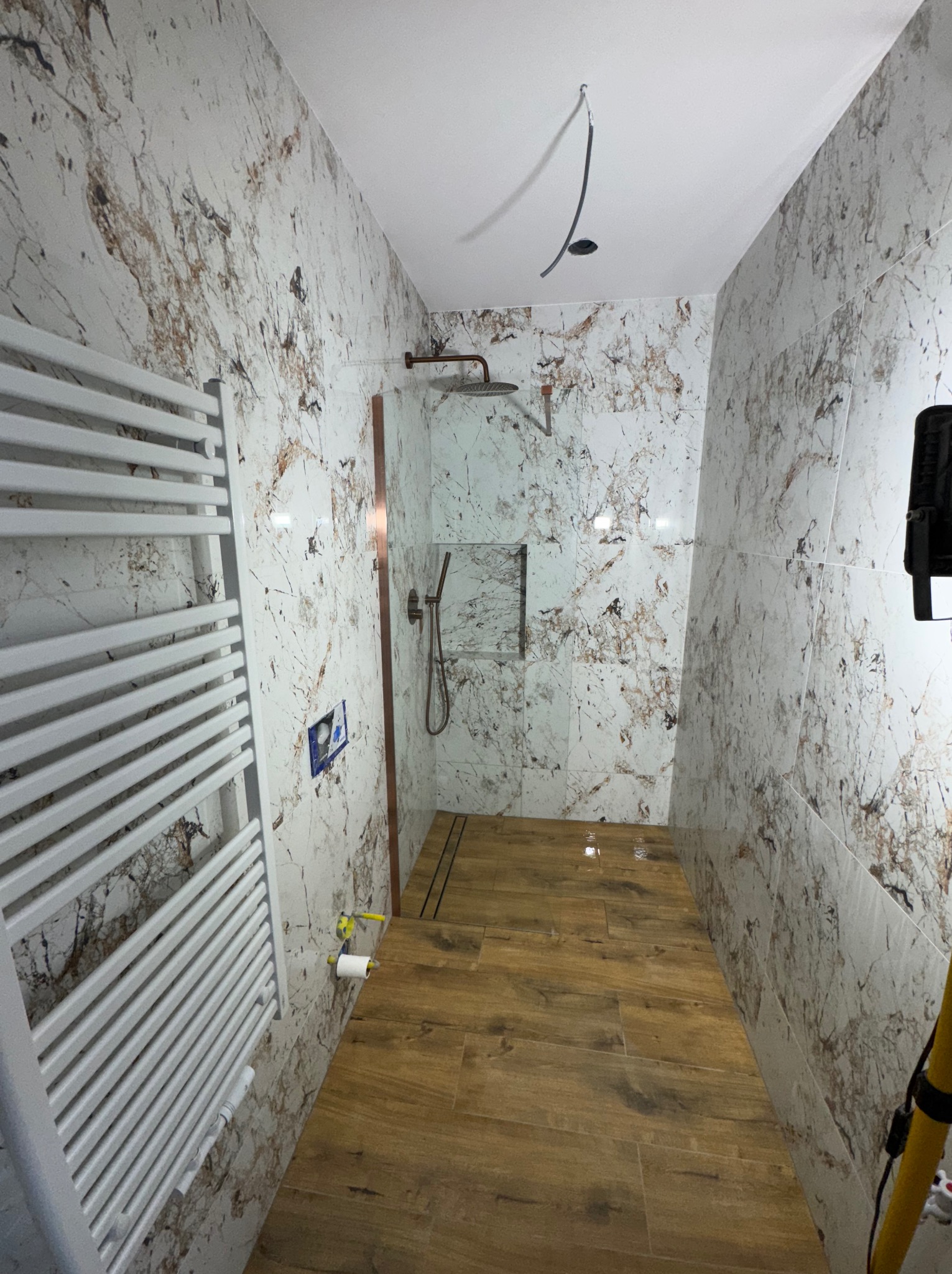 Nowoczesna łazienka z marmurowymi płytkami ściennymi i drewnopodobną podłogą. Widoczny grzejnik drabinkowy i kabina prysznicowa z deszczownicą. Minimalistyczny design.