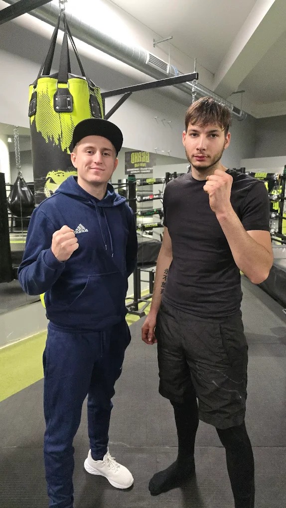 Dwóch mężczyzn w sportowym stroju pozuje w sali treningowej z workiem bokserskim w tle, prezentując gotowość do treningu.