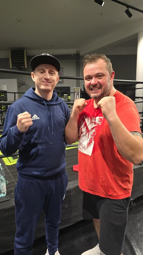 Dwóch mężczyzn w strojach sportowych pozuje z zaciśniętymi pięściami na tle ringu bokserskiego. Jeden w bluzie z kapturem, drugi w czerwonej koszulce.