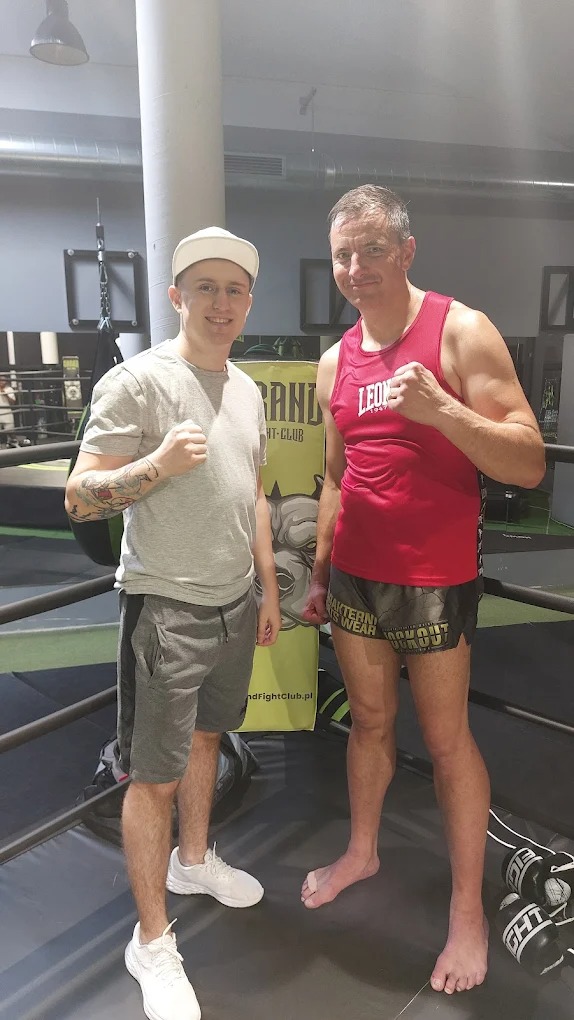 Dwóch mężczyzn w ringu bokserskim, jeden w stroju sportowym, drugi w casualowym ubraniu, obaj z zaciśniętymi pięściami, pozują do zdjęcia na tle logotypu klubu.