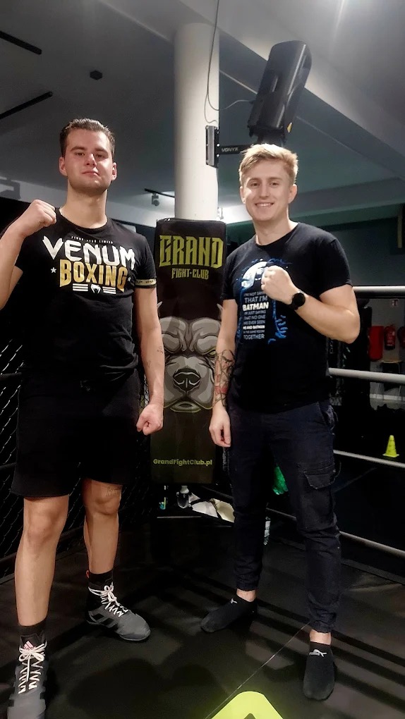 Dwóch mężczyzn w ringu bokserskim pozuje przed workiem treningowym Grand Fight Club, prezentując postawę gotową do walki. Jeden w koszulce Venum Boxing, drugi w koszulce z Batmanem.