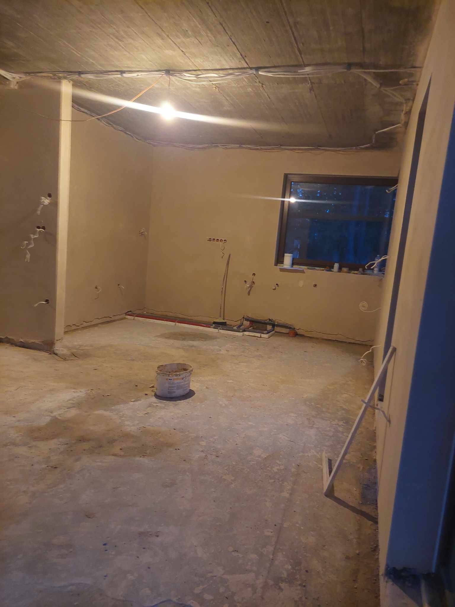 Pokój po nałożeniu tynków: gładkie, beżowe ściany, widoczne instalacje elektryczne i hydrauliczne, surowy betonowy sufit, wiadro po farbie na podłodze. Ujęcie z dołu.