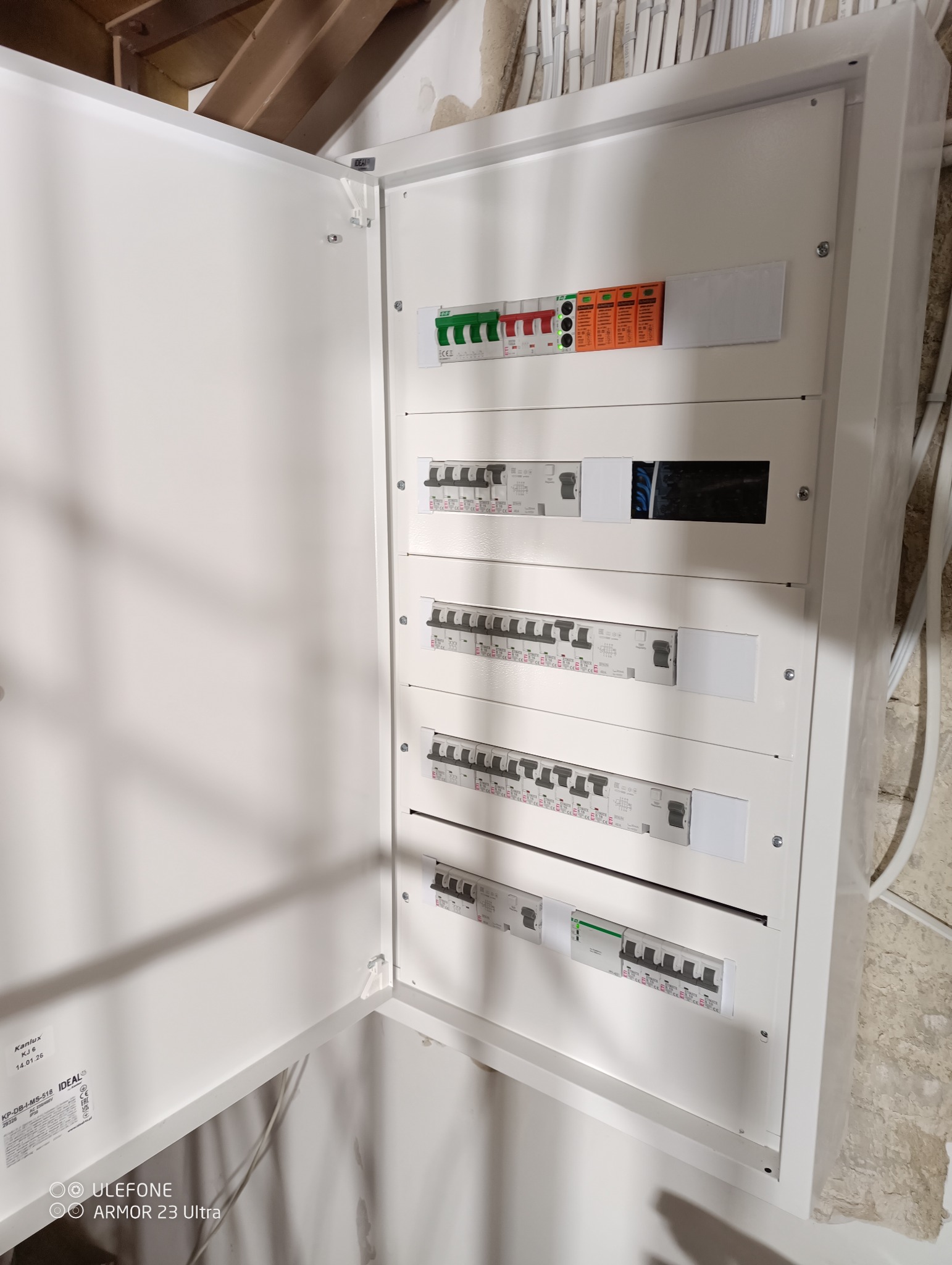 Otwarta, biała skrzynka rozdzielcza z bezpiecznikami i wyłącznikami różnicowoprądowymi, zamontowana na ścianie. Widoczne przewody elektryczne.