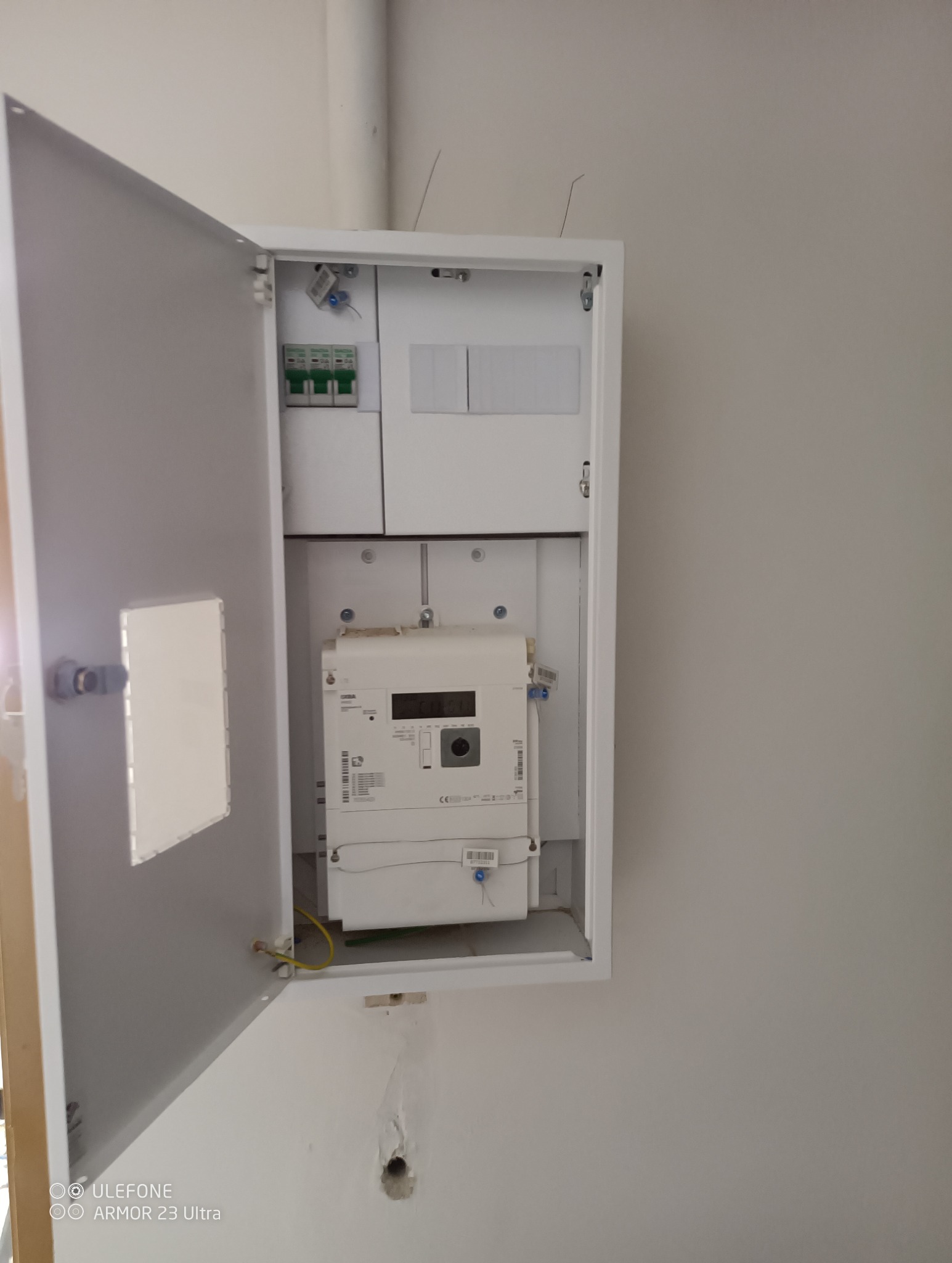 Otwarta skrzynka elektryczna z licznikiem i bezpiecznikami na ścianie. Widoczne przewody i elementy instalacji. Biała obudowa, jasne tło ściany.