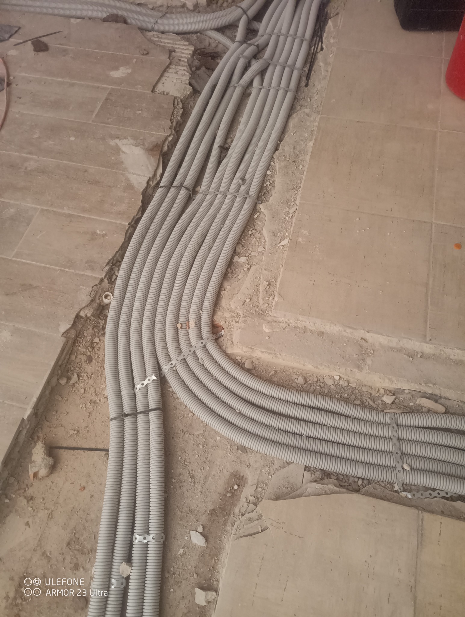 Szare rury karbowane ułożone w bruzdach podłogi, zabezpieczone uchwytami. Widoczne fragmenty płytek podłogowych i gruzu. Instalacja w trakcie prac.