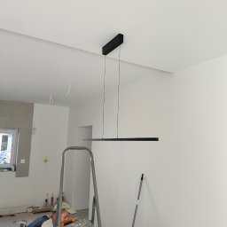 Sebastian Chmielewski - Monochromatyczna lampa LED zawieszona w jasnym wnętrzu. Elektryk na drabinie, w trakcie montażu. Widoczne narzędzia i elementy wykończeniowe. Minimalistyczny design.