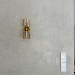 Sebastian Chmielewski - Ściana z dekoracyjną szarą fakturą, oświetlona złotym kinkietem z żarówką. Obok widoczny biały włącznik światła z czterema przyciskami i złotą ramką.