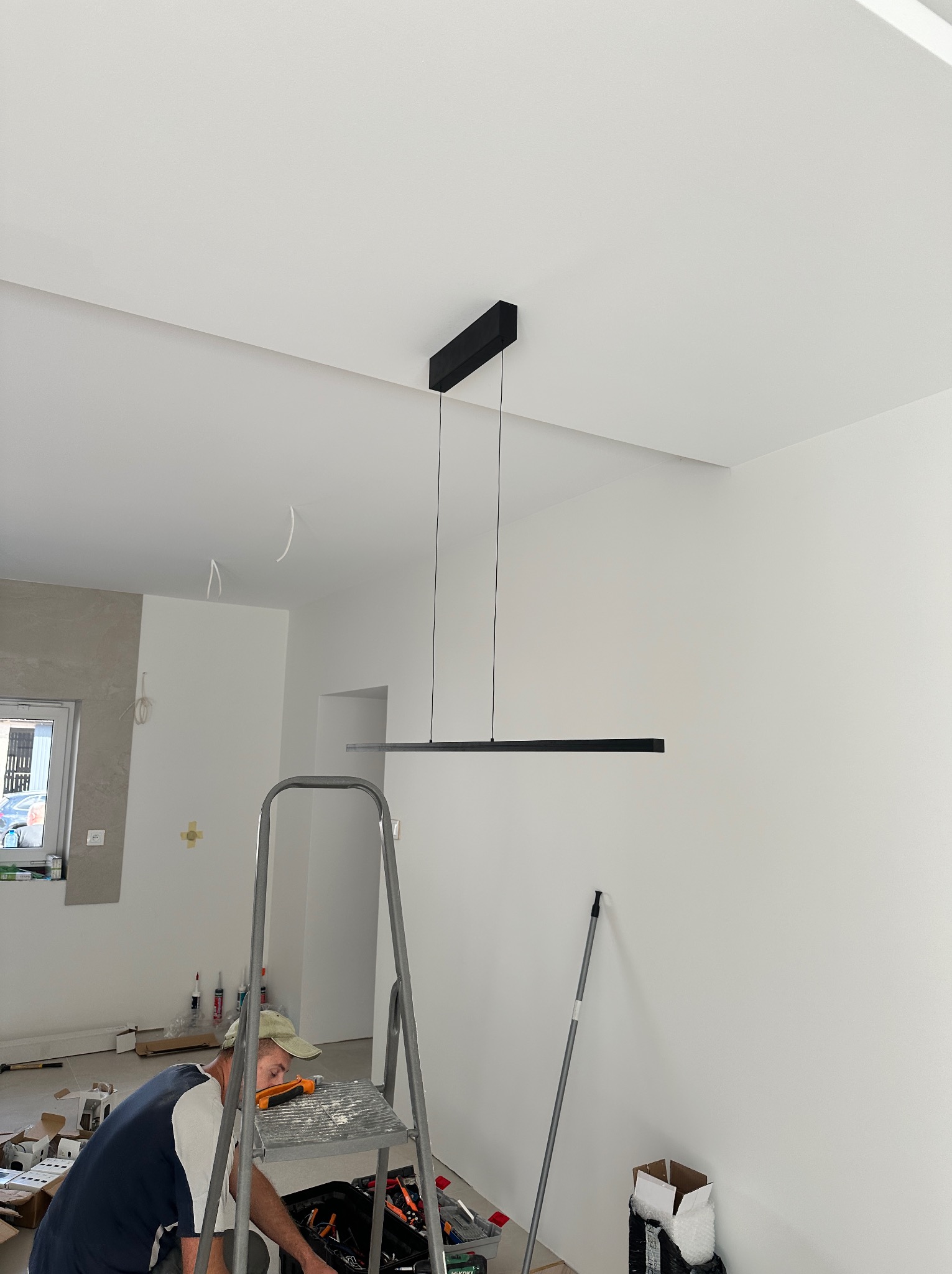 Monochromatyczna lampa LED zawieszona w jasnym wnętrzu. Elektryk na drabinie, w trakcie montażu. Widoczne narzędzia i elementy wykończeniowe. Minimalistyczny design.