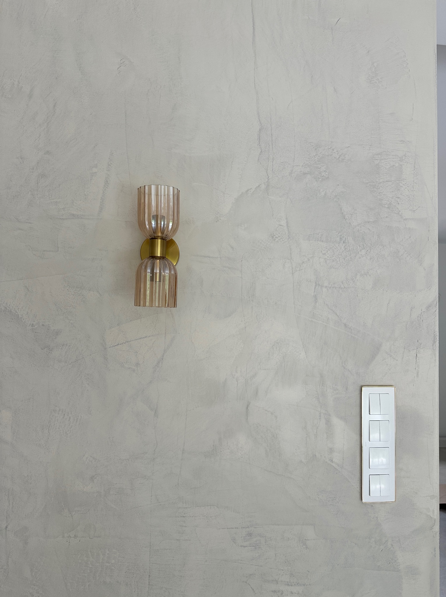 Ściana z dekoracyjną szarą fakturą, oświetlona złotym kinkietem z żarówką. Obok widoczny biały włącznik światła z czterema przyciskami i złotą ramką.