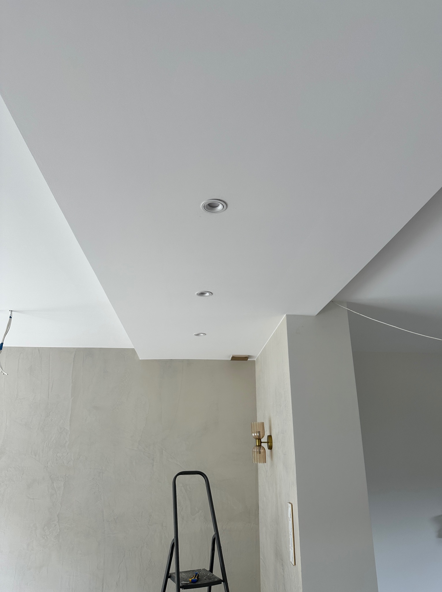 Wykończony sufit podwieszany z oświetleniem punktowym i ścianą z efektem dekoracyjnym. Widoczna drabina i lampa ścienna. Neutralna kolorystyka wnętrza.
