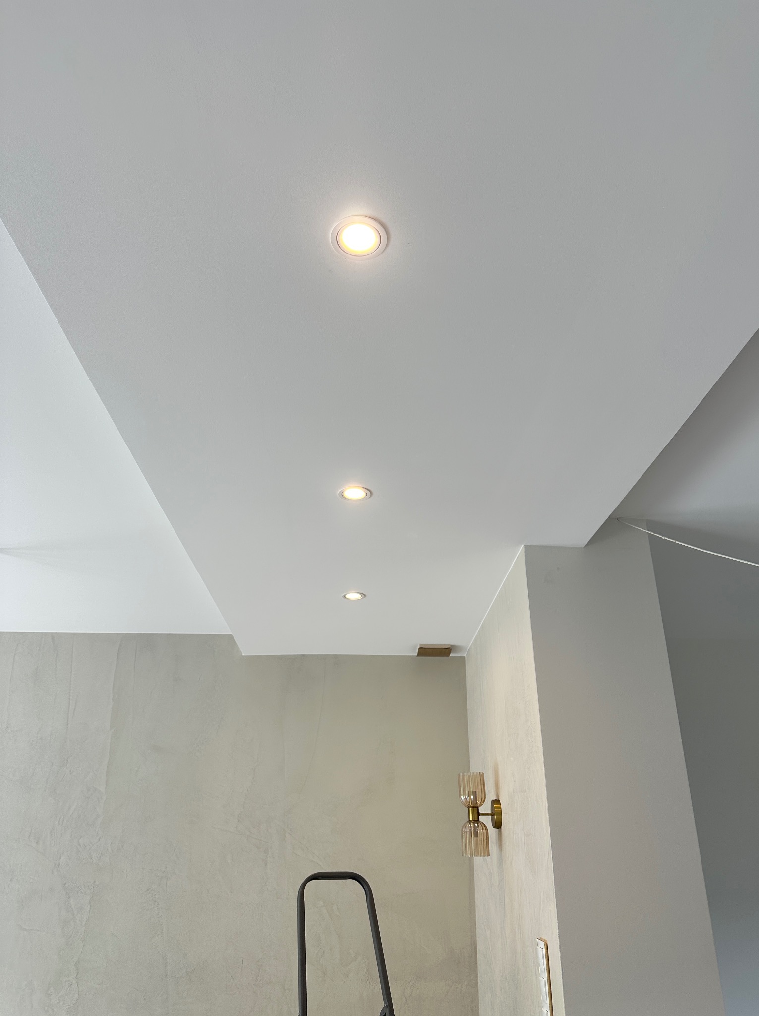 Biały sufit z trzema wpuszczonymi lampami LED w rzędzie, widok z dołu, fragment ściany z dekoracyjną lampą i drabina malarska w rogu. Minimalistyczne wykończenie wnętrza.