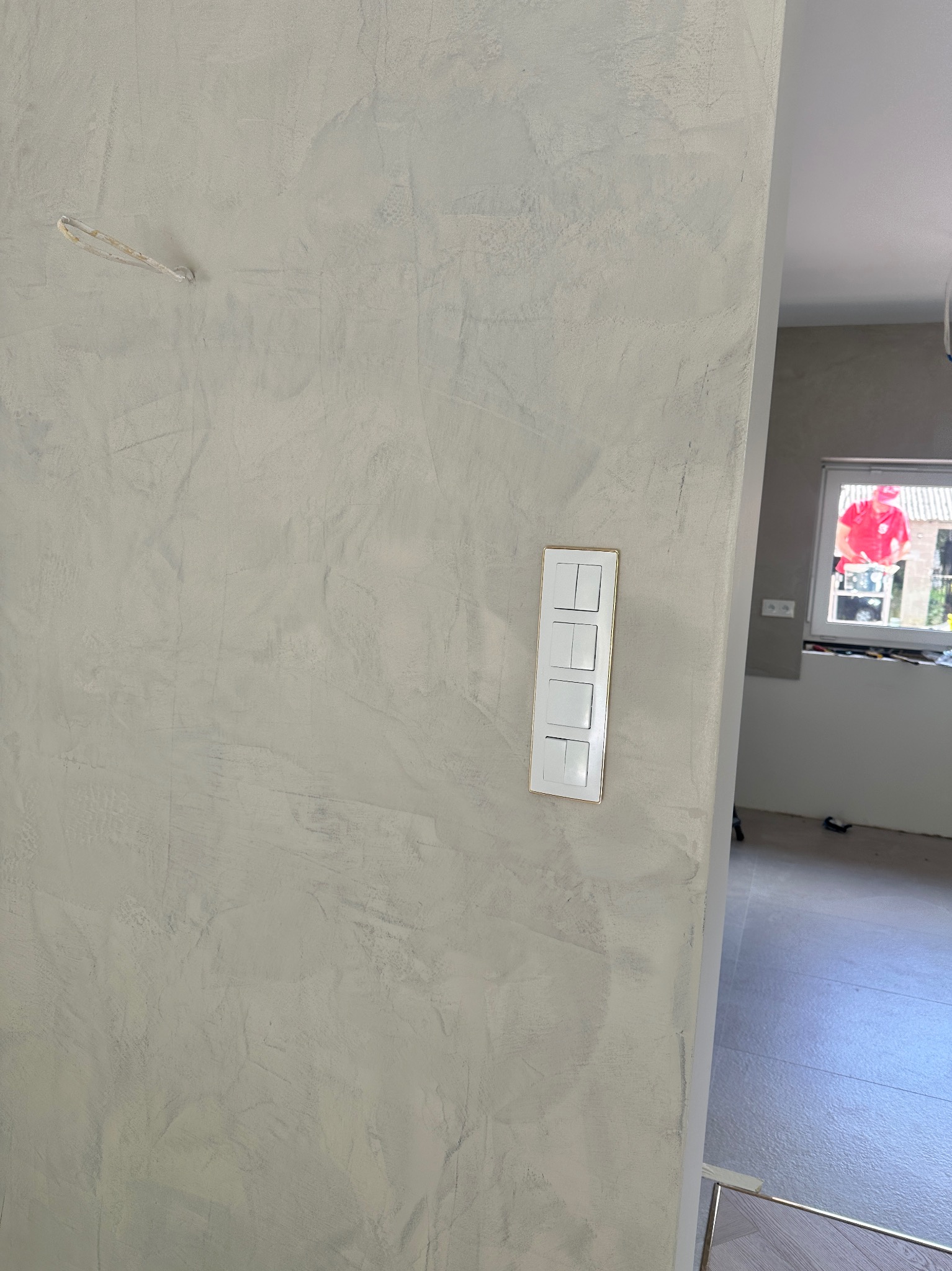 Ściana z dekoracyjnym szpachlowaniem w odcieniach szarości, z zamontowanym, białym panelem czterech włączników światła z delikatną, złotą ramką.