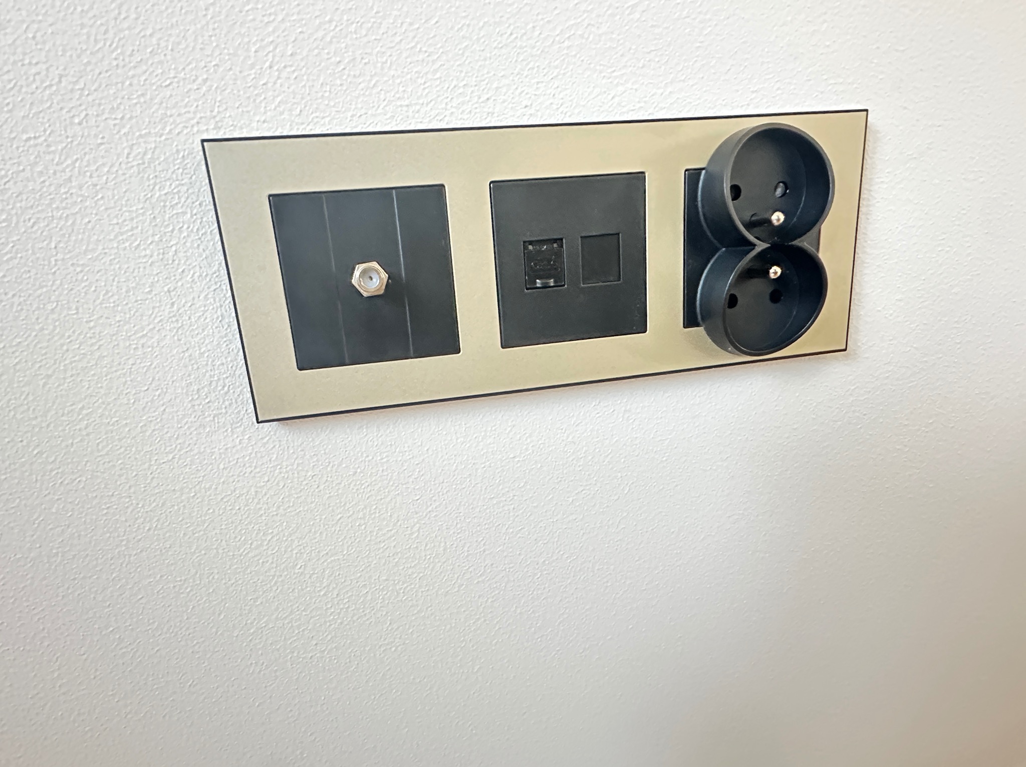 Panel z gniazdami: antenowe, internetowe i podwójne elektryczne, w czarnej obudowie z beżową ramką, zamontowany na białej ścianie. Nowoczesny design.
