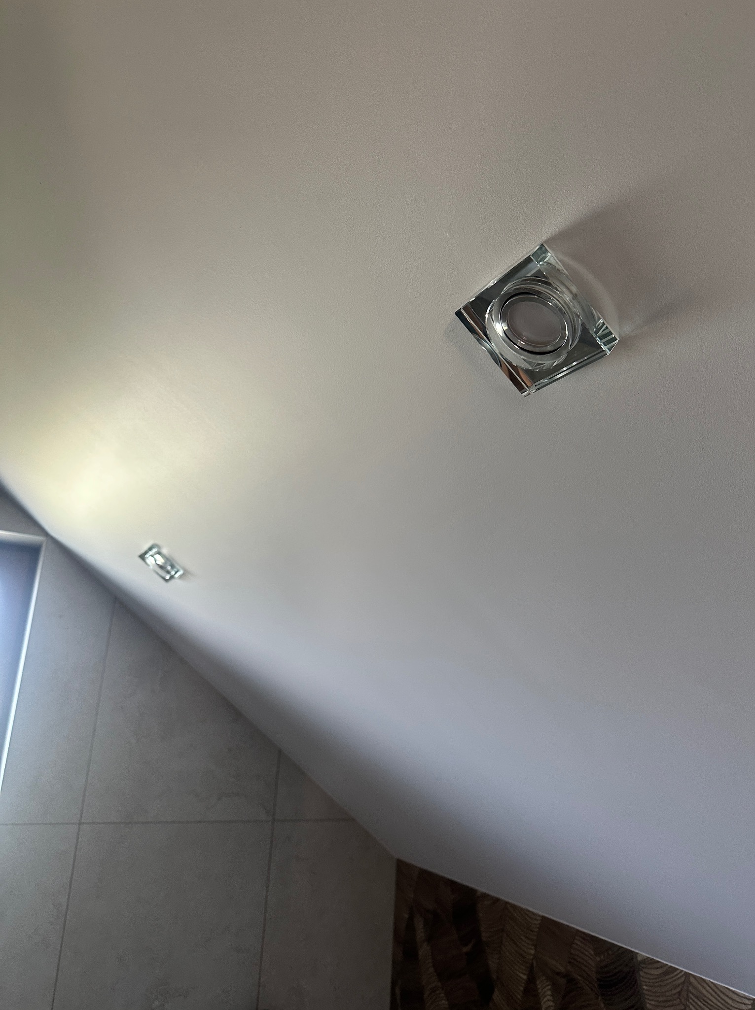 Biały sufit pod skosem z trzema kwadratowymi, szklanymi lampami halogenowymi. W tle fragment ściany z szarymi płytkami i dekoracyjną tapetą.