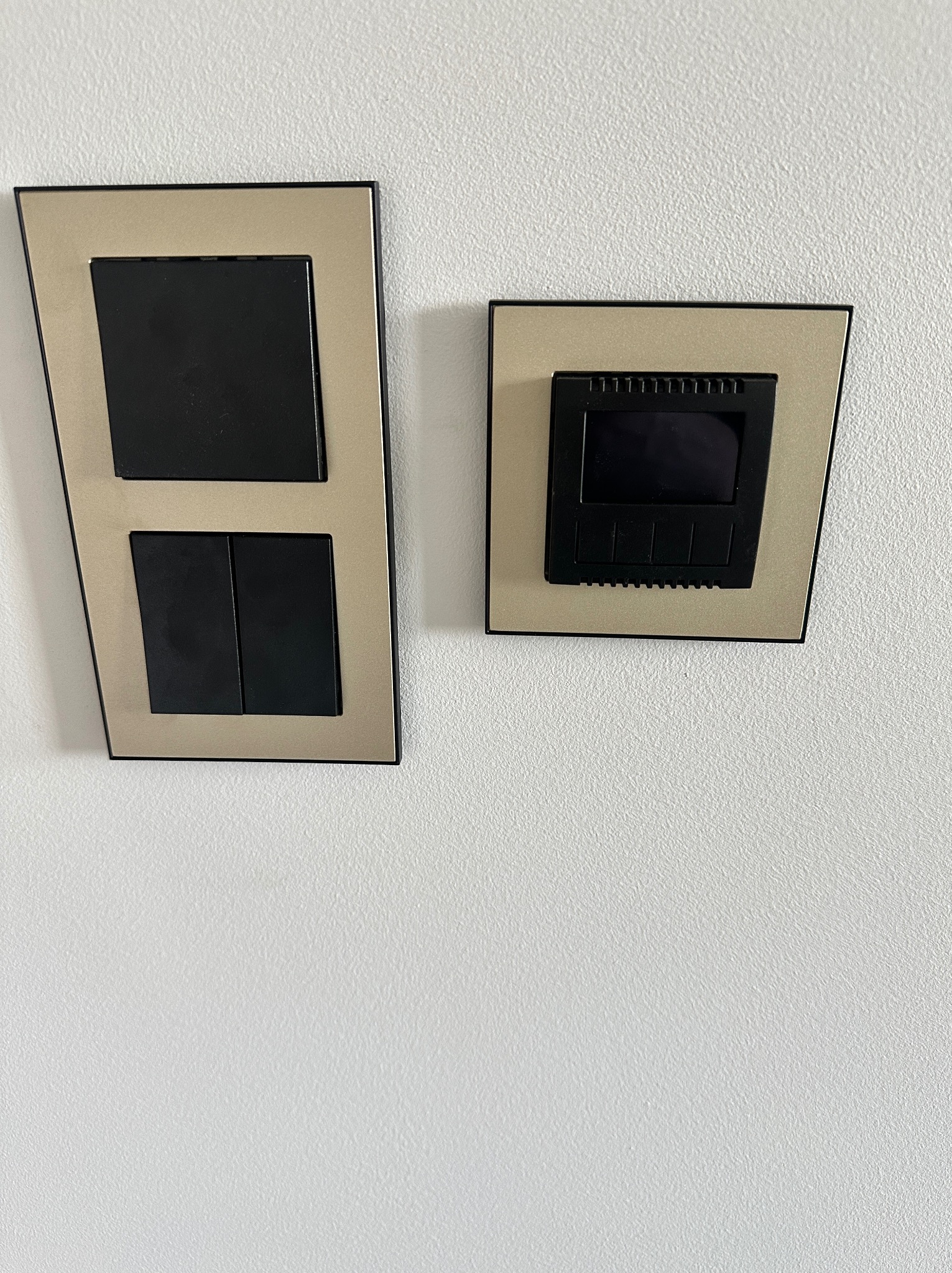 Dwa panele sterowania w złotych ramkach na białej ścianie w Lidzbarku. Jeden to włącznik światła, drugi to panel sterowania, prawdopodobnie ogrzewaniem.