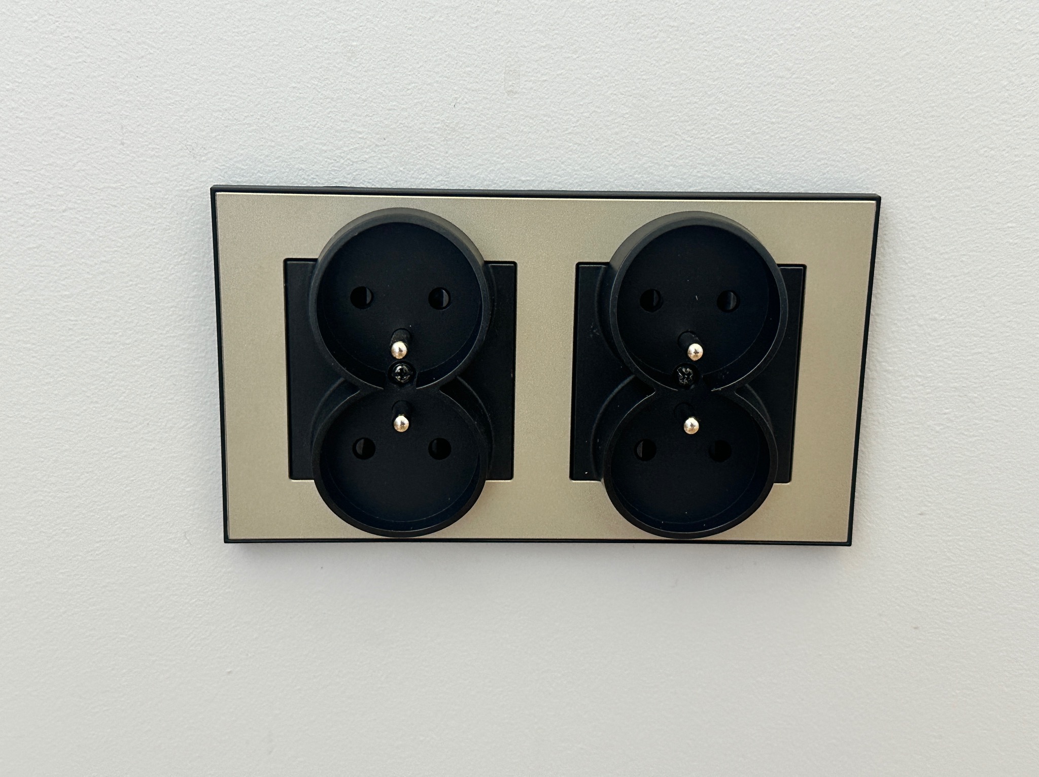 Dwa podwójne gniazdka elektryczne Schuko w srebrnej ramce, zamontowane na białej ścianie. Widok z bliska, oświetlenie dzienne.