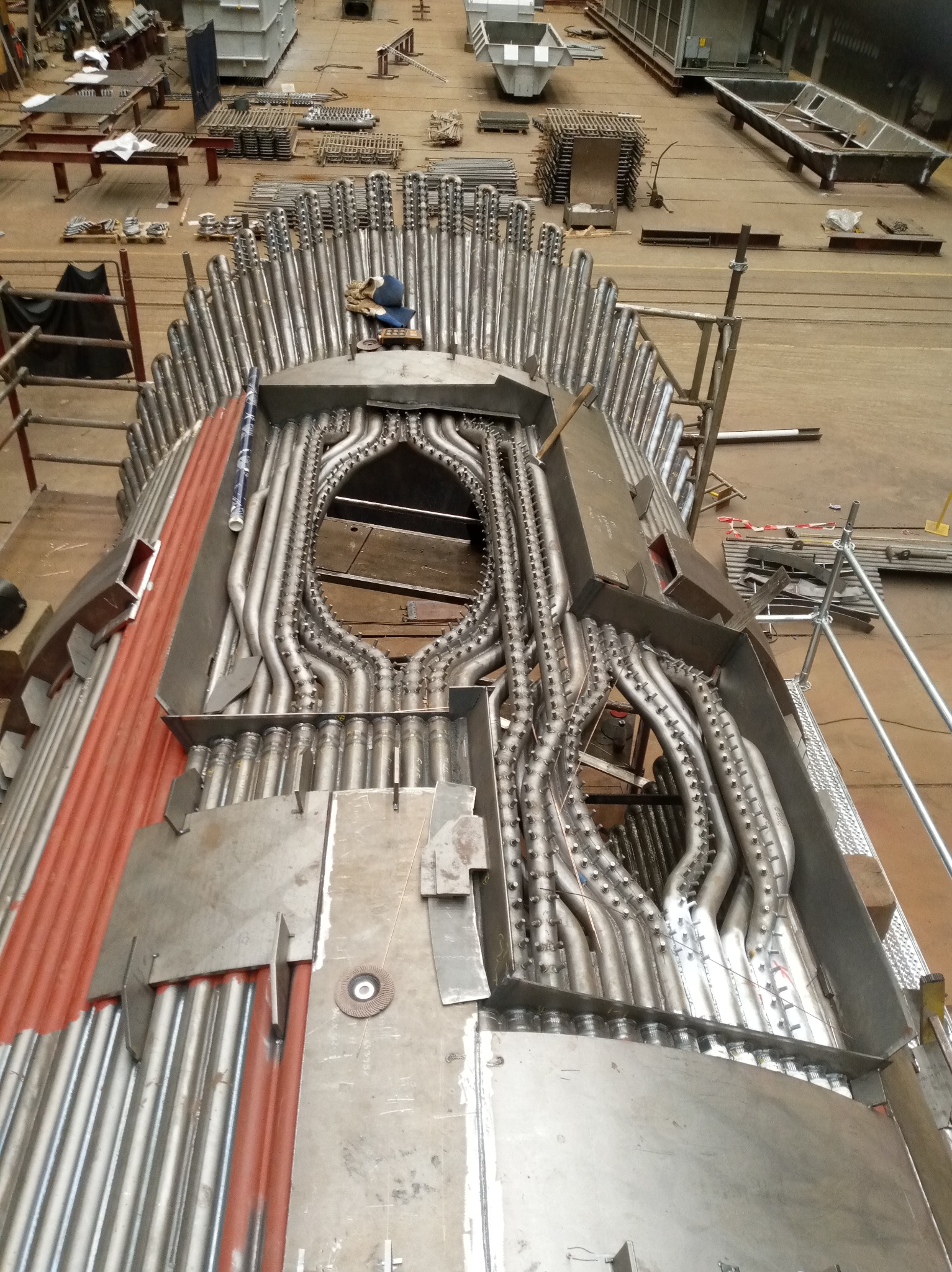 Konstrukcja kotła przemysłowego w trakcie budowy. Widoczne liczne rury, elementy metalowe, rusztowania i narzędzia. Ujęcie z góry, podkreślające złożoność instalacji.