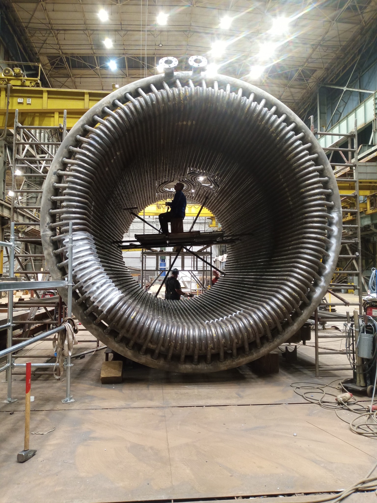 Obróbka metalu: spawanie spiralnej konstrukcji rurowej w hali przemysłowej. Dwóch spawaczy pracuje wewnątrz i na zewnątrz konstrukcji, widoczne rusztowania i narzędzia.