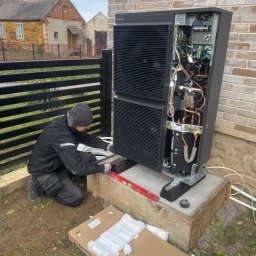 BLUETHERMX Adrian Pietrzyk - Instalator poziomuje pompę ciepła Panasonic na betonowym fundamencie. Widoczne otwarte wnętrze urządzenia i elementy instalacji. W tle budynek z cegły.