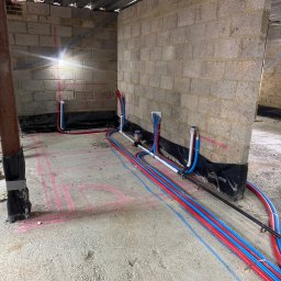 BLUETHERMX Adrian Pietrzyk - Instalacja rur grzewczych (czerwone i niebieskie) w surowym wnętrzu budynku z betonową podłogą i ścianami z pustaków. Widoczne oznaczenia instalacji na podłodze.