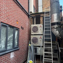 BLUETHERMX Adrian Pietrzyk - Dwa agregaty klimatyzacyjne Mitsubishi zamontowane na zewnątrz budynku z cegły, obok metalowej drabiny i rur wentylacyjnych. Widoczne kable i ostrzeżenie o wysokim napięciu.