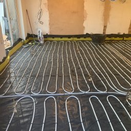 BLUETHERMX Adrian Pietrzyk - Białe rury ogrzewania podłogowego ułożone na czarnej folii z żółtymi brzegami, widoczne uchwyty mocujące i fragment ściany w trakcie remontu.