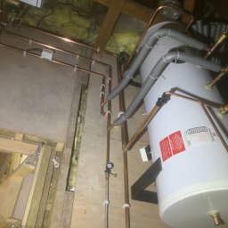 BLUETHERMX Adrian Pietrzyk - Instalacja bojlera w przestrzeni poddasza z widocznymi miedzianymi rurami, izolacją termiczną i elementami mocującymi. Widoczne drewniane elementy konstrukcyjne.