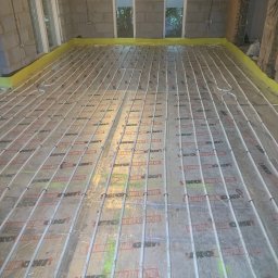 BLUETHERMX Adrian Pietrzyk - Ułożone rury ogrzewania podłogowego na folii izolacyjnej w surowym wnętrzu. Widoczne okna i fragment ściany z bloczków. Żółta taśma brzegowa.