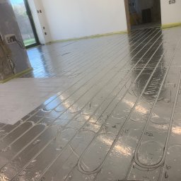 BluethermX - Błyszcząca instalacja ogrzewania podłogowego z rurkami w kształcie pętli, widoczne oznaczenia numeracji, żółta taśma na obrzeżach, surowy filar w tle.
