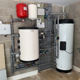 BLUETHERMX Adrian Pietrzyk - Dwa białe zasobniki Vaillant, czerwony zbiornik wyrównawczy i szare rury izolowane na tle ściany z betonu komórkowego. Instalacja w kotłowni.