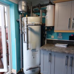 BLUETHERMX Adrian Pietrzyk - Daikin: Biały zasobnik ciepłej wody użytkowej z szarymi rurami w izolacji, obok szare szafki kuchenne i turkusowa ściana. Widoczny fragment drzwi balkonowych.