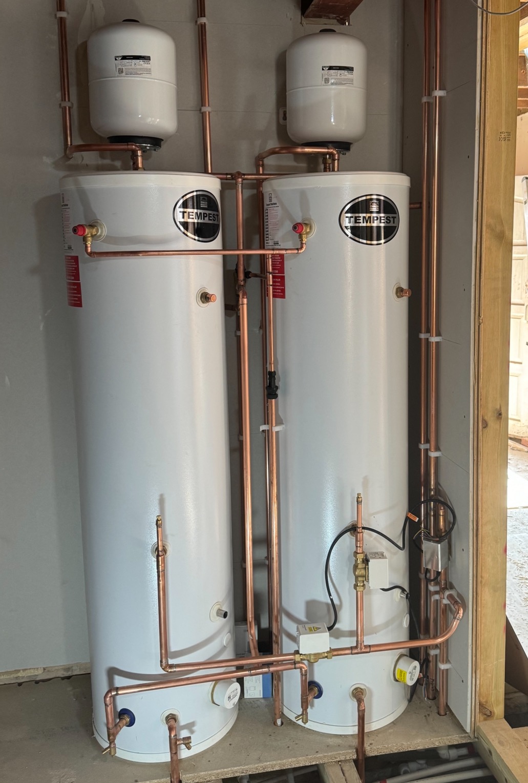 Dwa białe, pionowe zbiorniki Tempest z miedzianymi rurami i białymi zbiornikami wyrównawczymi, zamontowane w pomieszczeniu z surowymi ścianami i podłogą.