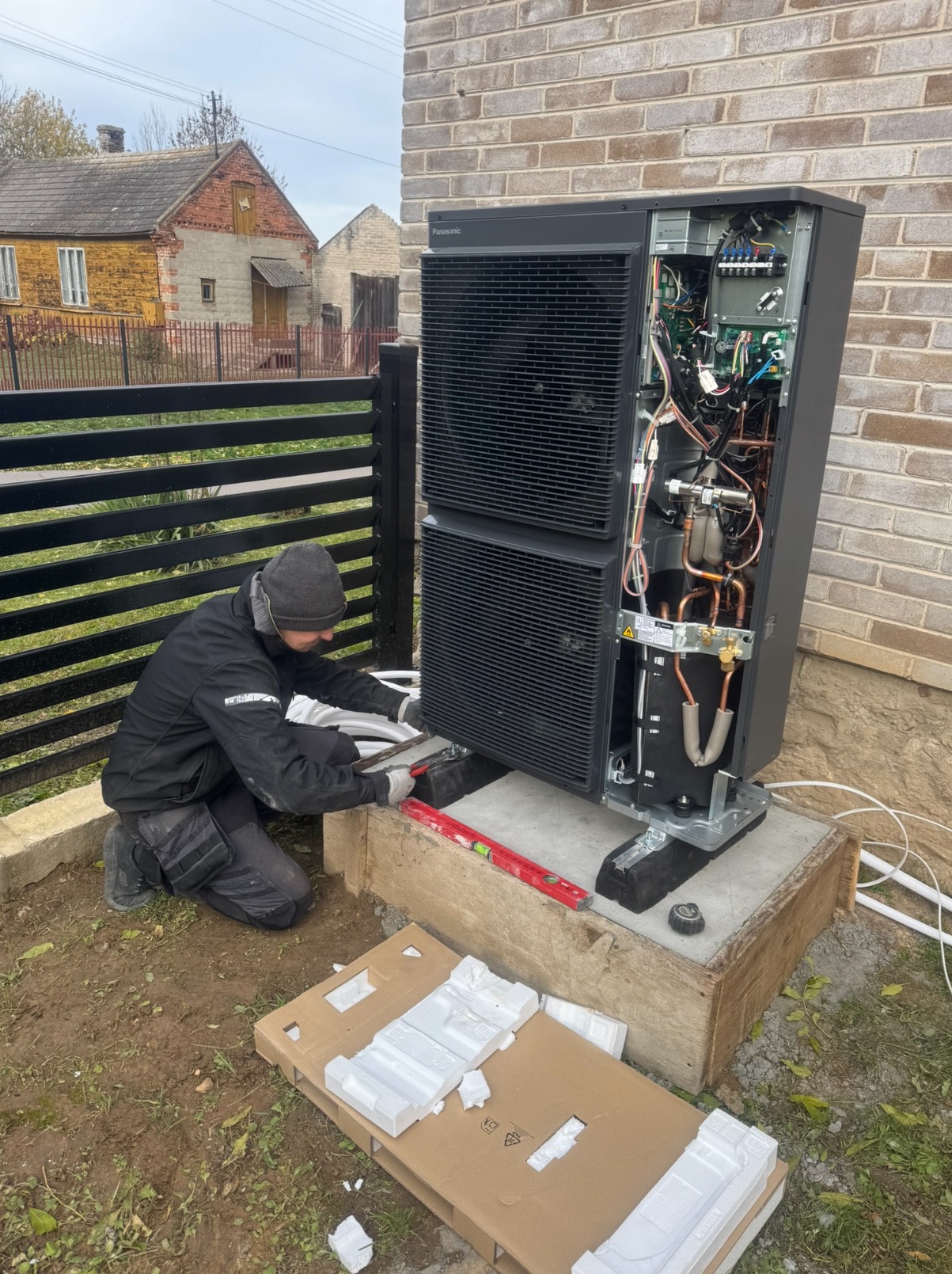 Instalator poziomuje pompę ciepła Panasonic na betonowym fundamencie. Widoczne otwarte wnętrze urządzenia i elementy instalacji. W tle budynek z cegły.