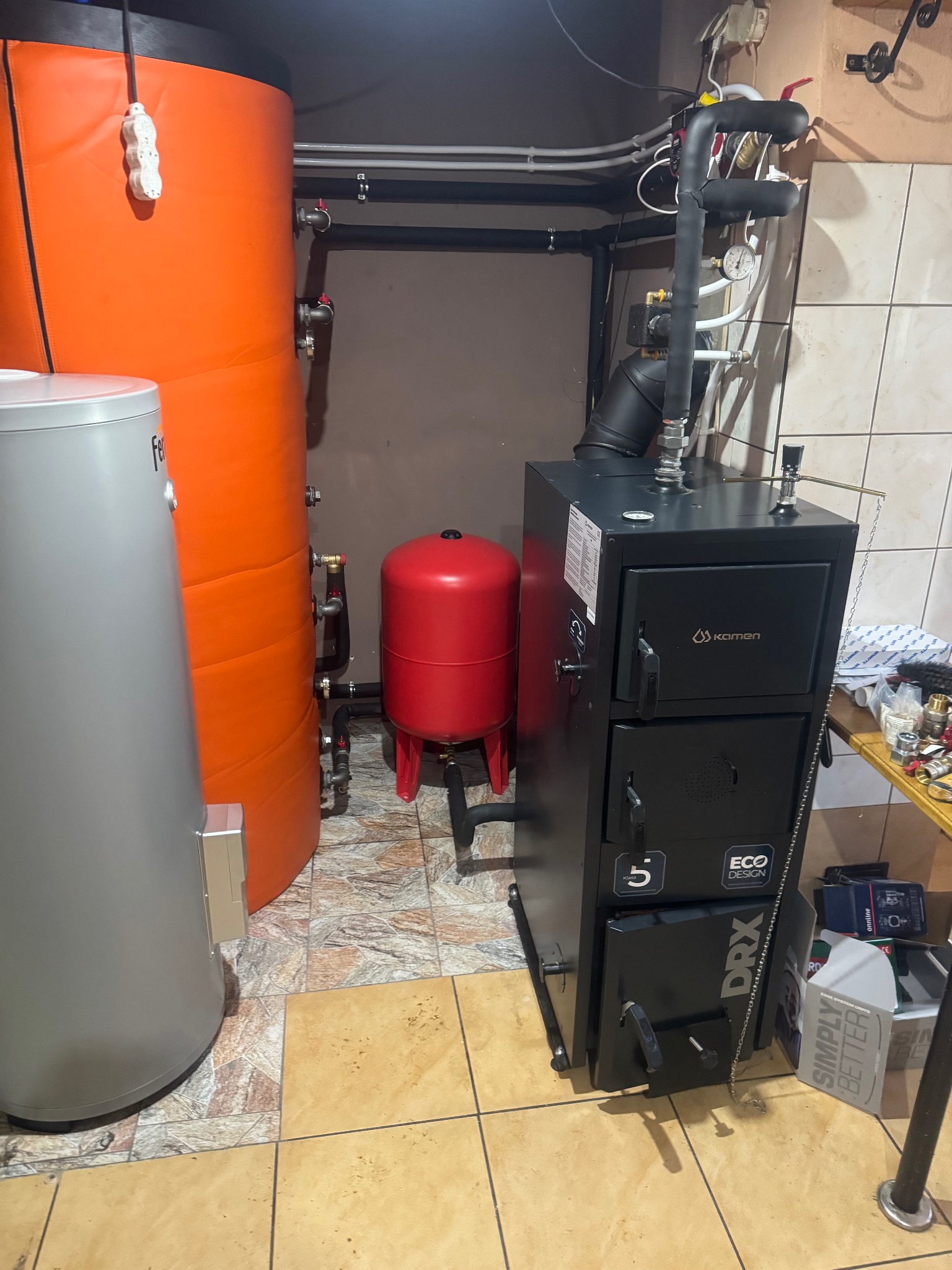 Czarny piec CO marki Kamen Eco Design DRX, obok zbiorniki w kolorze szarym i pomarańczowym oraz czerwony zbiornik wyrównawczy. Instalacja w kotłowni.