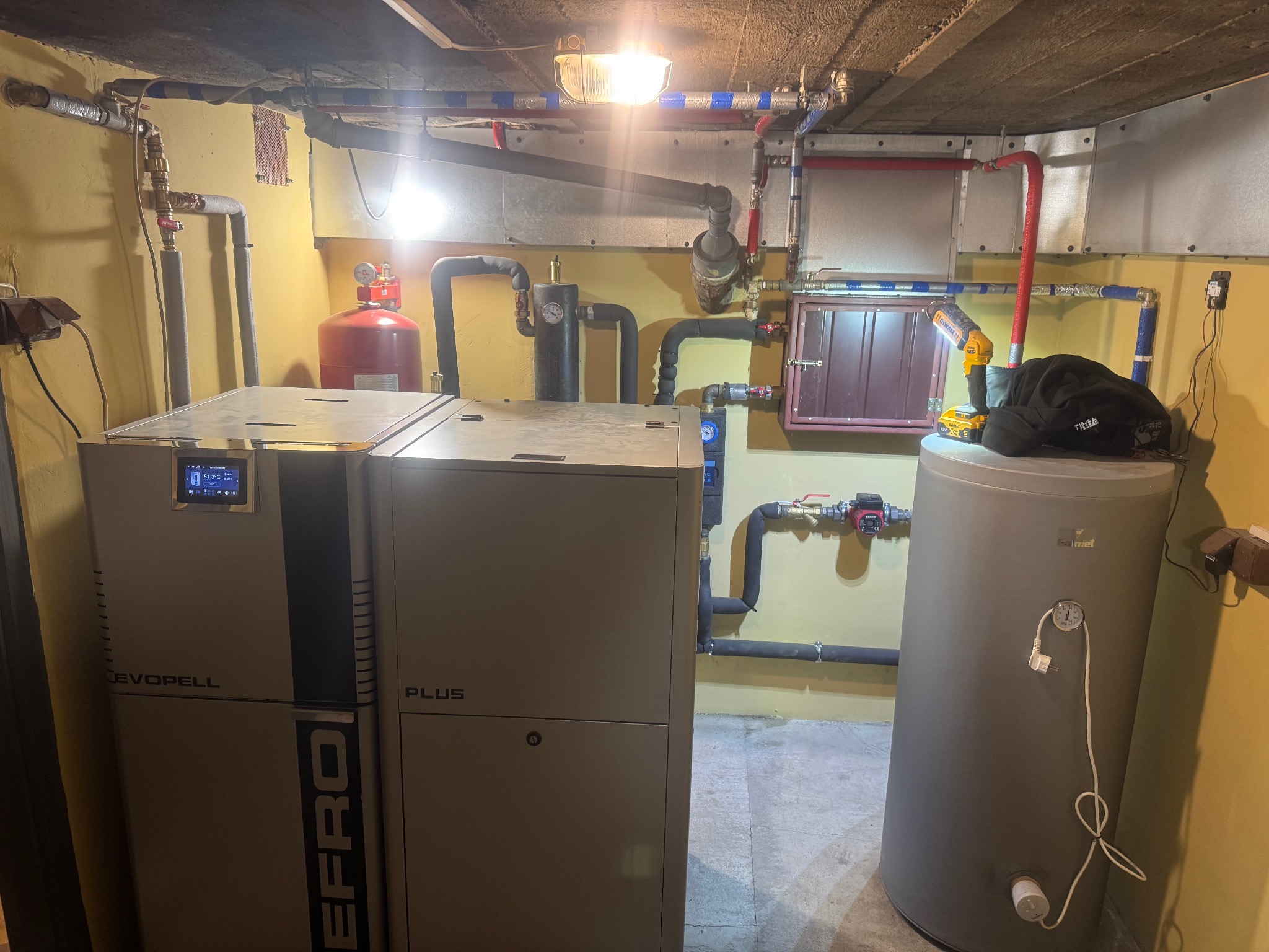 Zestaw urządzeń grzewczych: piec marki EFROPELL PLUS i zasobnik Calmet, połączone rurami. Widoczne elementy instalacji CO i CWU, narzędzia na zasobniku. Żółte ściany, sufit z instalacjami.