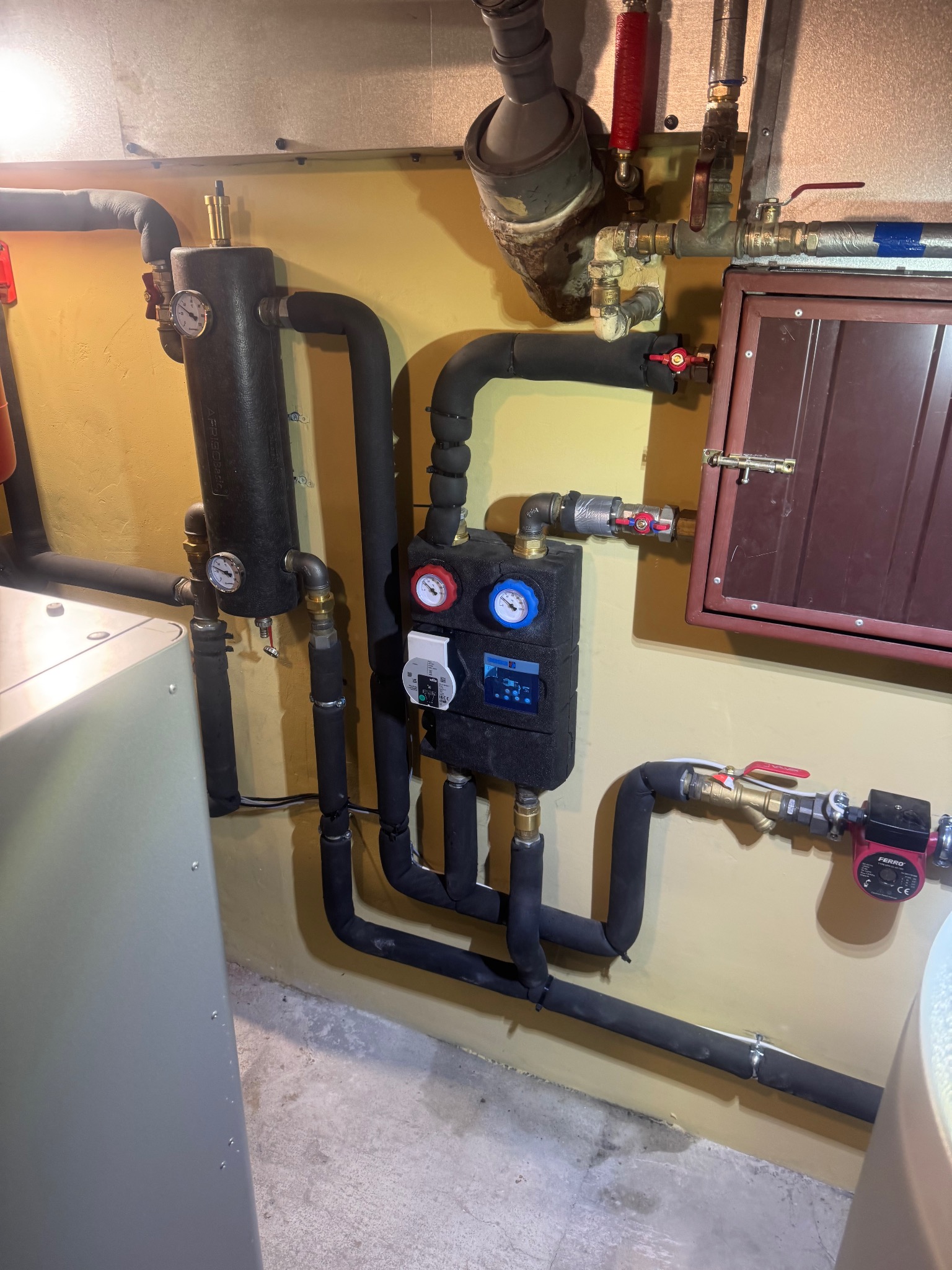 Złożona instalacja grzewcza z czarnymi rurami izolowanymi, manometrami, zaworami i sterownikiem, zamontowana na żółtej ścianie w kotłowni. Widoczne elementy pompy obiegowej.