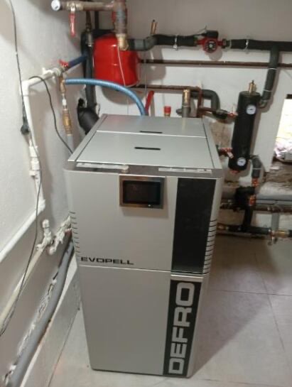 Szary piec marki DEFRO EVOPELL z podłączonymi rurami w kotłowni. Widoczny zbiornik wyrównawczy i elementy instalacji grzewczej.