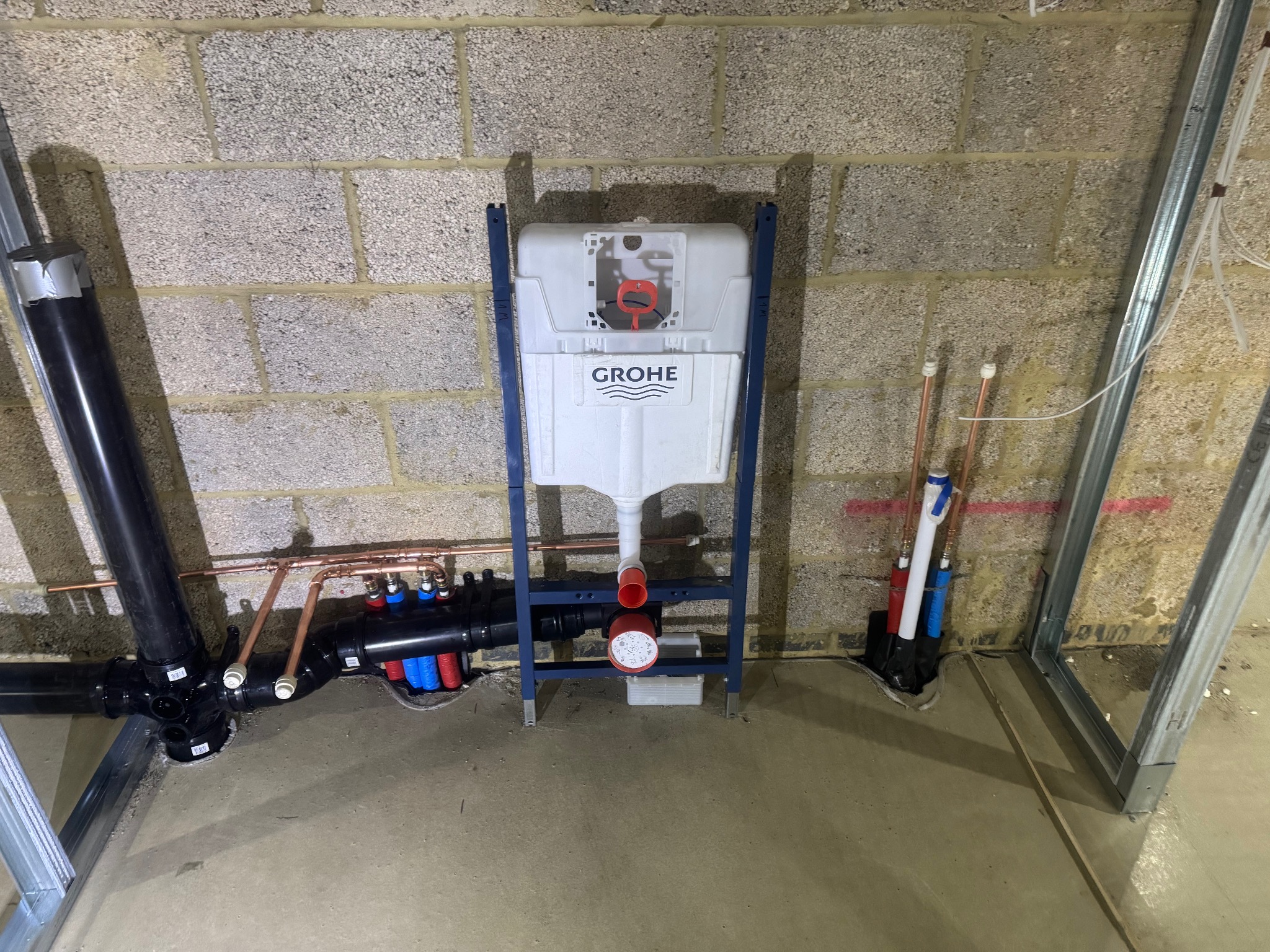 Stelaż podtynkowy Grohe z instalacją wodno-kanalizacyjną na tle surowej ściany z betonu komórkowego i metalowych profili. Widoczne rury miedziane i plastikowe.