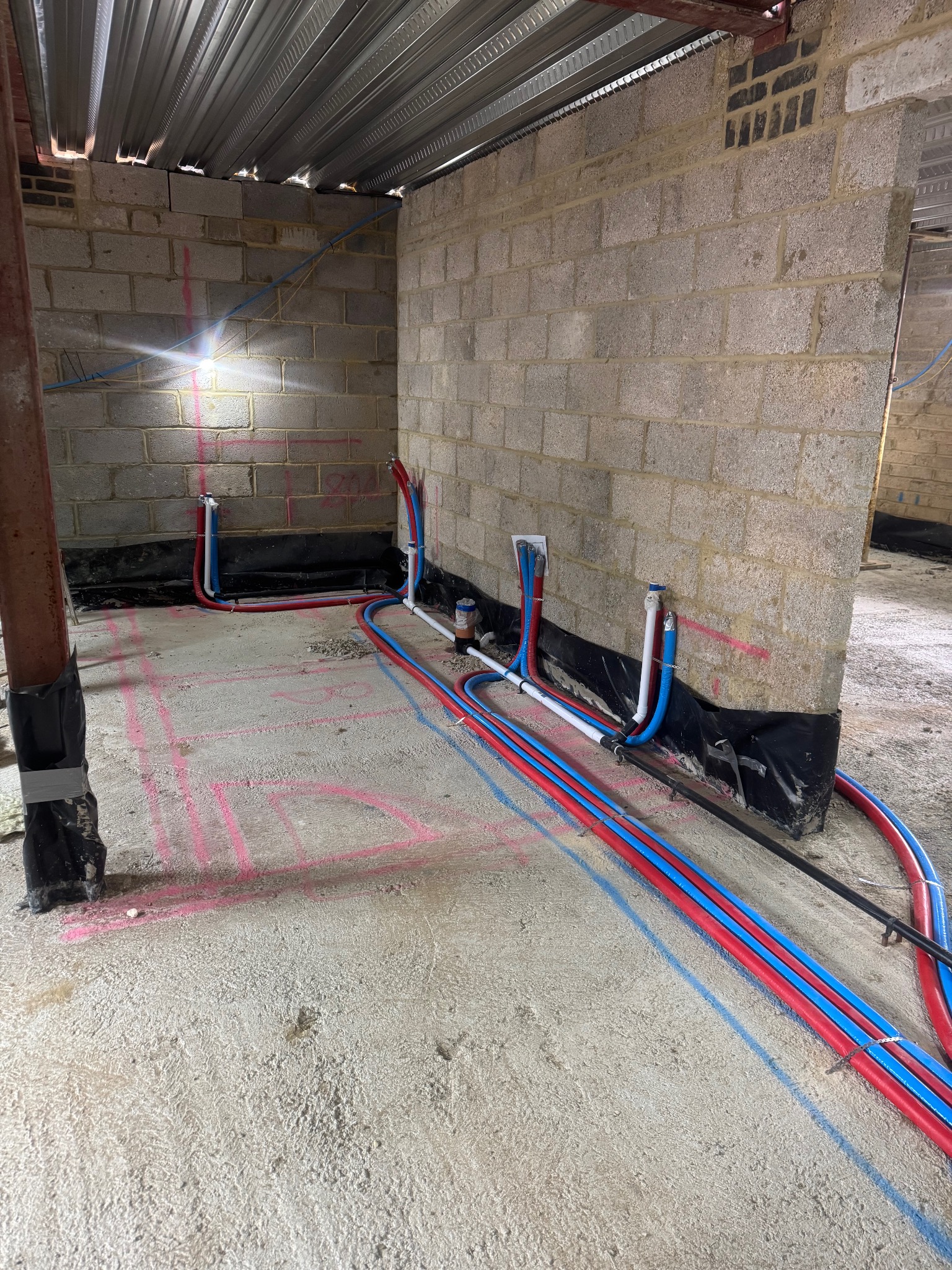 Instalacja rur grzewczych (czerwone i niebieskie) w surowym wnętrzu budynku z betonową podłogą i ścianami z pustaków. Widoczne oznaczenia instalacji na podłodze.