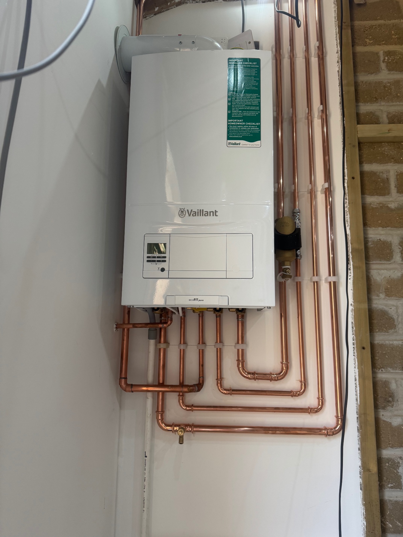 Biały piec gazowy Vaillant ecoFIT pure z miedzianą instalacją, zamontowany na białej ścianie, z widocznymi rurami i elementami mocującymi. Ujęcie z bliska, z boku.